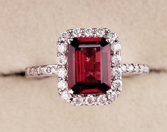 May include: A red gemstone ring with a halo of smaller diamonds. The ring is set in white metal.