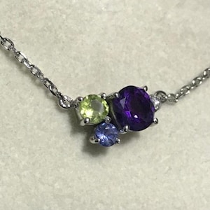 May include: A silver chain necklace with a cluster of three gemstones. The largest gemstone is a deep purple amethyst, surrounded by a smaller light green peridot and a tiny blue topaz.