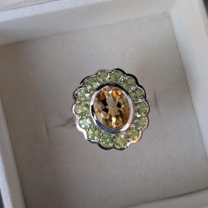 Könnte beinhalten: Ein silberner Ring mit einem großen ovalen Topas-Edelstein, der von einem Heiligenschein aus kleineren grünen Peridot-Edelsteinen umgeben ist.