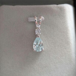 May include: A silver pendant featuring a pear-shaped, light blue gemstone and a smaller, round, clear stone. The pendant hangs from a silver bail, set against a gray background.