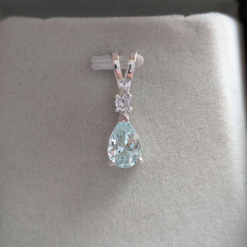 May include: A silver pendant featuring a pear-shaped, light blue gemstone and a smaller, round, clear stone. The pendant hangs from a silver bail, set against a gray background.