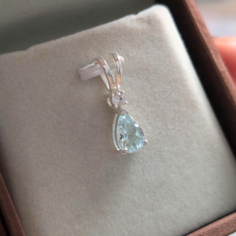 May include: A silver pendant with a pear-shaped, light blue gemstone. The pendant has a small, round pink gemstone above the larger blue stone. The pendant is displayed on a beige surface.