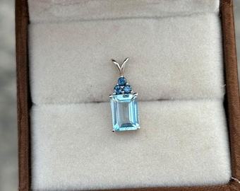Könnte beinhalten: Ein Anhänger mit einem hellblauen Edelstein im quadratischen Smaragdschliff, umgeben von kleineren blauen Edelsteinen. Der Anhänger ist in Silber gefasst.