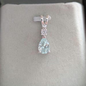 May include: A silver pendant with a pear-shaped, light blue gemstone and a small, round, clear stone above it. The pendant has a V-shaped bail for a chain. The background is a light gray, textured surface.