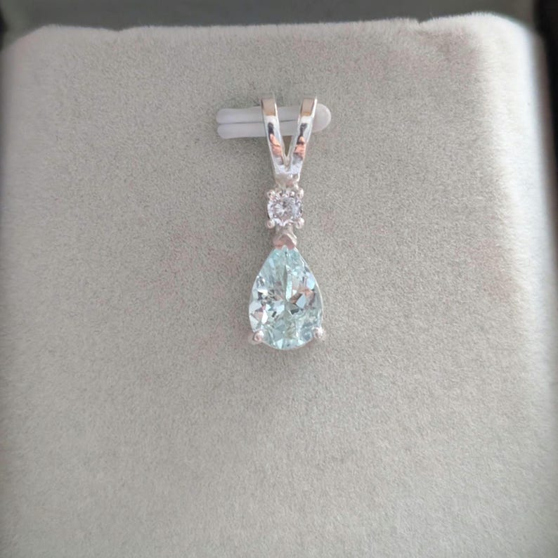 May include: A silver pendant with a pear-shaped, light blue gemstone and a small, round, clear stone above it. The pendant has a V-shaped bail for a chain. The background is a light gray, textured surface.