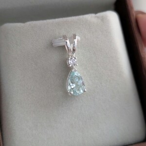 May include: A silver pendant with a pear-shaped, light blue gemstone and a small, round, clear stone above it. The pendant is displayed on a gray surface inside a brown box.