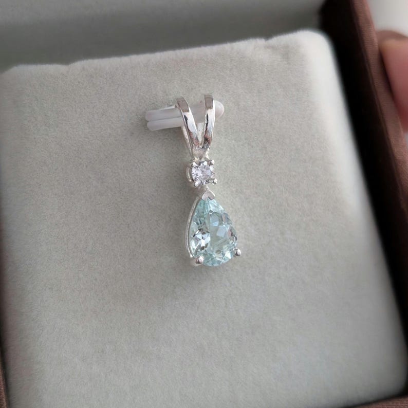 May include: A silver pendant with a pear-shaped, light blue gemstone and a small, round, clear stone above it. The pendant is displayed on a gray surface inside a brown box.