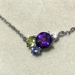 May include: A silver chain necklace with a cluster of gemstones. The center stone is a large, round, purple amethyst. The other stones are a smaller, round, green peridot and a small, round, blue sapphire.