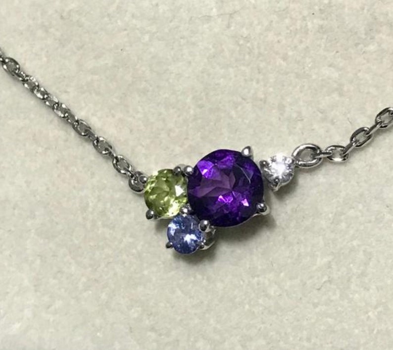 May include: A silver chain necklace with a cluster of gemstones. The center stone is a large, round, purple amethyst. The other stones are a smaller, round, green peridot and a small, round, blue sapphire.