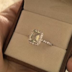 May include: An emerald-cut engagement ring with a halo of smaller stones and a band of pave diamonds. The ring is set in a brown ring box with a beige interior. The ring is a symbol of love and commitment.