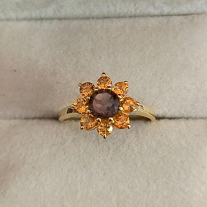 May include: A gold ring with a brown gemstone surrounded by smaller orange gemstones in a flower shape.