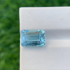 Puede incluir: Una gema rectangular de color azul claro con una superficie facetada. La gema está colocada sobre un fondo blanco con un fondo verde borroso. La gema tiene una apariencia clara y transparente.