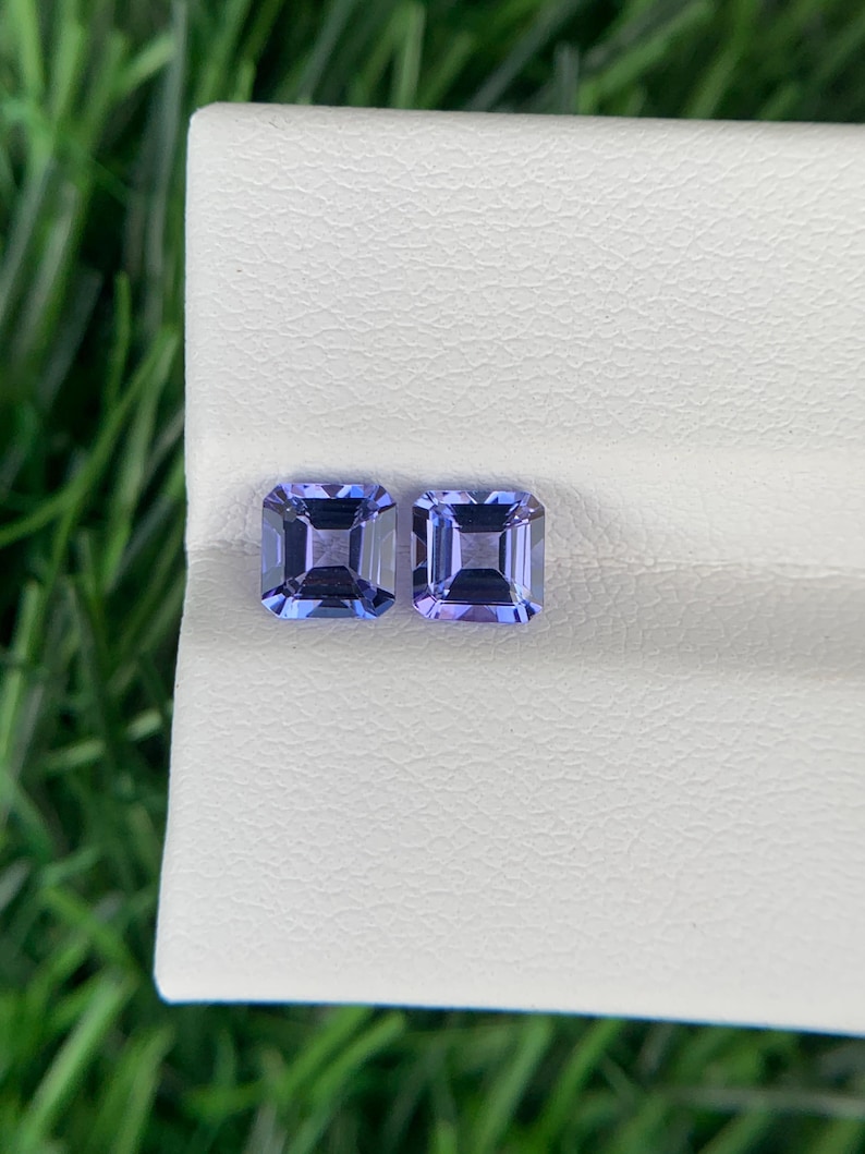 May include: Two square-cut, light blue gemstones with a slight purple hue. The gems are displayed on a white surface, with green grass visible in the background. The gemstones are likely for jewelry.