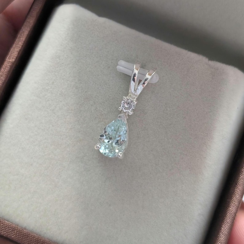 May include: A silver pendant with a pear-shaped, light blue gemstone. The pendant has a small, round gemstone above the larger stone and is displayed in a brown jewelry box.