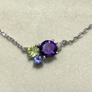 May include: A silver chain necklace with a cluster of three gemstones. The largest gemstone is a round, purple amethyst. The other two gemstones are smaller and round, one is green and one is blue.