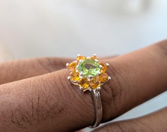 Puede incluir: Un anillo de plata con una piedra preciosa verde rodeada de piedras preciosas amarillas más pequeñas.