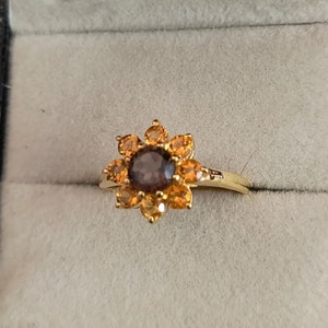 May include: A gold ring with a brown gemstone surrounded by smaller orange gemstones in a flower shape.