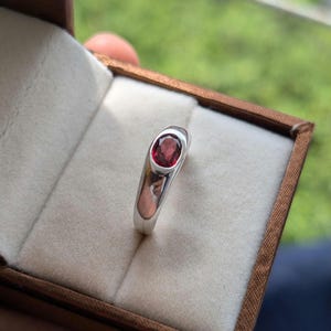 May include: A silver ring featuring a deep red oval gemstone, set in a polished silver band. The ring is presented in a brown jewellery box with a cream-coloured interior. The gemstone is the central feature, reflecting light.