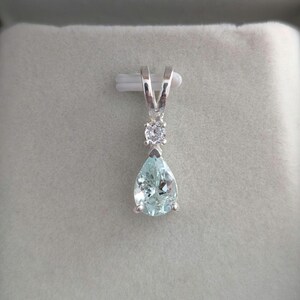 May include: A silver pendant with a pear-shaped, light blue gemstone. A small, round, clear stone sits above the larger stone. The pendant hangs from a silver bail. The gemstone has a faceted appearance.
