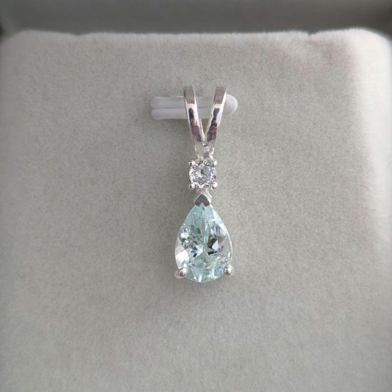 May include: A silver pendant with a pear-shaped, light blue gemstone. A small, round, clear stone sits above the larger stone. The pendant hangs from a silver bail. The gemstone has a faceted appearance.