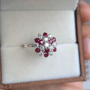 May include: A silver ring with a floral design, set with red and white gemstones. The ring is presented in a neutral-toned jewellery box.