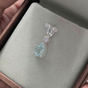 May include: A silver pendant with a pear-shaped, light blue gemstone and a small, round, clear stone. The pendant is set in a silver setting and is displayed in a brown jewelry box.