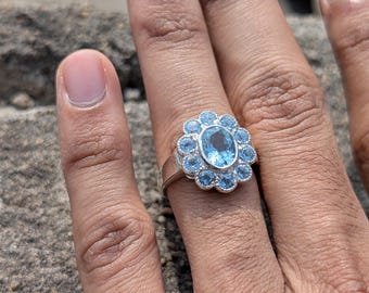 May include: A silver ring featuring a large oval-cut, light blue gemstone surrounded by smaller, round, light blue gemstones. The ring's design resembles a flower. The ring is on a person's finger.