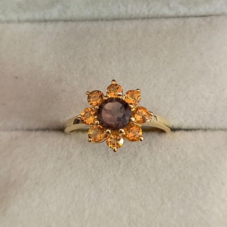 May include: A gold ring with a brown gemstone surrounded by a cluster of orange gemstones in a flower shape.