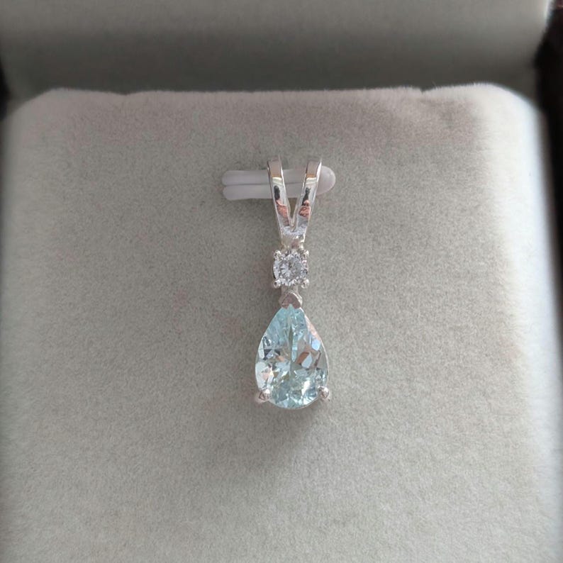 May include: A silver pendant with a pear-shaped, light blue gemstone and a small, round, clear stone. The pendant has a V-shaped bail and is set against a gray background.