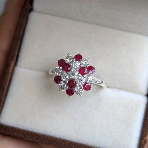May include: A silver ring featuring a cluster of round, deep red gemstones interspersed with smaller, clear gemstones. The ring's design resembles a floral arrangement, set against a neutral background.