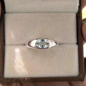 May include: A silver ring featuring a single, oval-cut, light blue gemstone set in a smooth, polished metal band. The ring is centred within a neutral-toned, padded jewellery box, showcasing the reflective surface of the metal and the clarity of the stone.