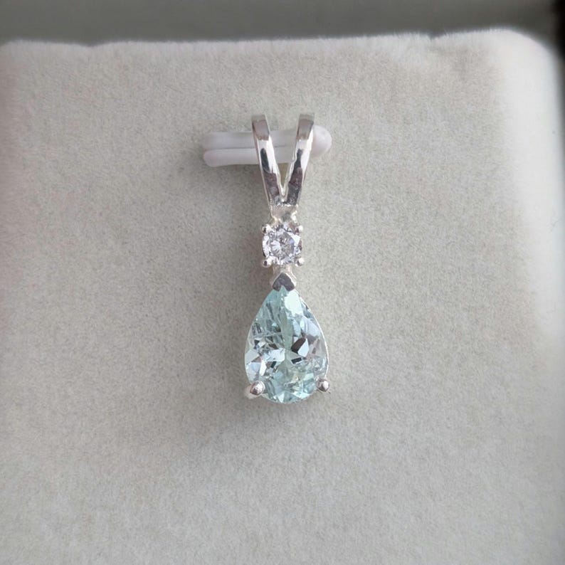 May include: A silver pendant featuring a pear-shaped, light blue gemstone. Above the main stone is a small, round, clear gemstone, and the pendant has a V-shaped bail. The pendant is set against a light gray background.