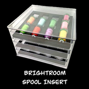 May include: A clear acrylic Brightroom Spool Insert with multiple drawers, each holding colorful spools of thread. The text "BRIGHTROOM SPOOL INSERT" is displayed at the bottom of the image. The spools are in shades of green, orange, pink, and purple.
