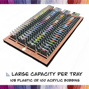 May include: A wooden tray with multiple rows of clear plastic bobbins, each labeled with a number. The tray is designed to hold 108 plastic or 100 acrylic bobbins. The text "LARGE CAPACITY PER TRAY" and "108 PLASTIC OR 100 ACRYLIC BOBBINS" is visible.
