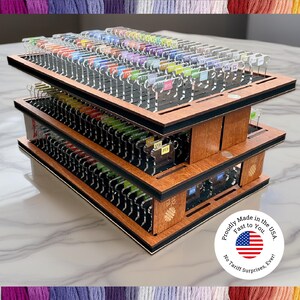 May include: A three-tiered wooden organizer with multiple slots, holding a variety of colorful thread spools. The organizer has a dark border and is labeled with small tags. The product is proudly made in the USA.