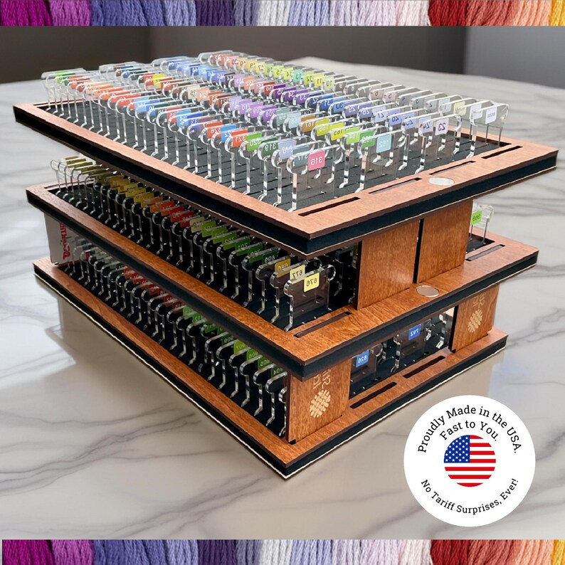 May include: A three-tiered wooden organizer with multiple slots, holding a variety of colorful thread spools. The organizer has a dark border and is labeled with small tags. The product is proudly made in the USA.
