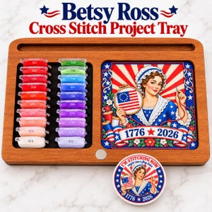 May include: A wooden cross stitch project tray featuring a rectangular inset with a colorful illustration of a woman holding an American flag hoop, text reading 1776 and 2026, and two columns of numbered embroidery floss bobbins. Includes a round magnet.