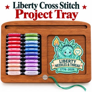 May include: A rectangular wooden project tray featuring a slot for embroidery floss bobbins, organized in two columns of red, pink, green, blue, and purple threads. A sticker depicts the Statue of Liberty with the text Liberty Needles & Thread 1776-2026.