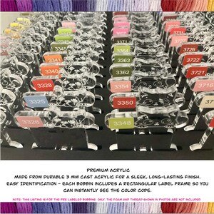 May include: Clear acrylic bobbins with color-coded labels, arranged in rows. Each bobbin has a cat-shaped design and a rectangular label frame. The text "PREMIUM ACRYLIC" and other details are visible. The bobbins are designed for easy color code identification.