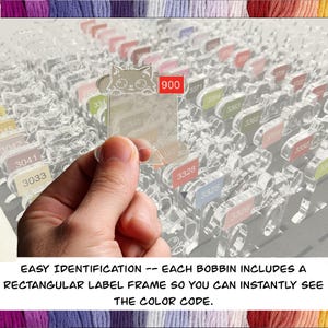 May include: A clear acrylic bobbin holder with multiple slots, each labeled with a number. A hand holds a clear acrylic label with a cat design and the number 900. The text "EASY IDENTIFICATION -- EACH BOBBIN INCLUDES A RECTANGULAR LABEL FRAME SO YOU CAN INSTANTLY SEE THE COLOR CODE."