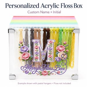 May include: A clear acrylic floss box featuring a large letter M decorated with floral embroidery patterns and the text Mary's Floss Box. The box includes a row of pastel-colored hangers at the top for organizing embroidery thread skeins.