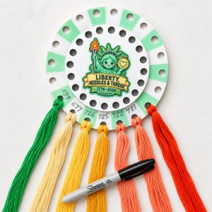 May include: A circular white and mint green embroidery floss organizer featuring a central illustration of the Statue of Liberty holding a needle and embroidery hoop with the text Liberty, Needles & Thread 1776-2026, with six colored threads and a black Sharpie.