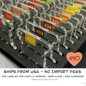 May include: Clear acrylic bobbins with color-coded labels, arranged in rows on a black foam insert. Each bobbin has a number on a colored label. The image includes the number 490 in a heart-shaped label.