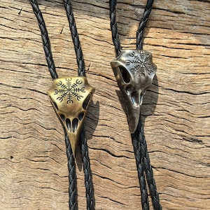 May include: Two bolo ties with raven skull pendants. One is bronze-colored, the other is silver-colored. Both have black braided cords. The pendants feature intricate designs, including a symbol at the top. The background is a wooden surface.
