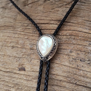 May include: A black leather bolo tie with a silver and white pearlescent pendant. The pendant has a decorative filigree design.