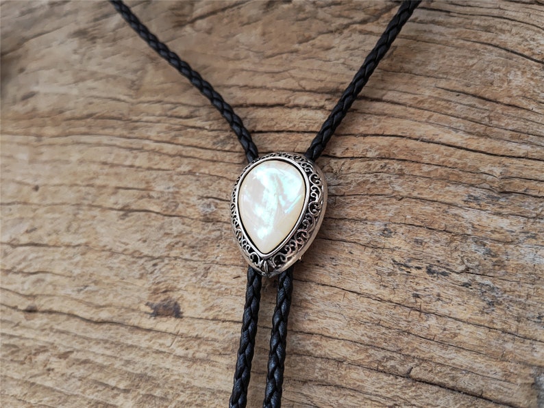 May include: A black leather bolo tie with a silver and white pearlescent pendant. The pendant has a decorative filigree design.