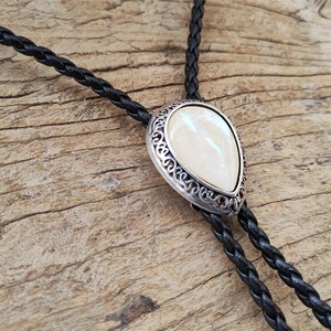 May include: A black braided bolo tie with a silver-toned pendant featuring a white pearlescent stone.