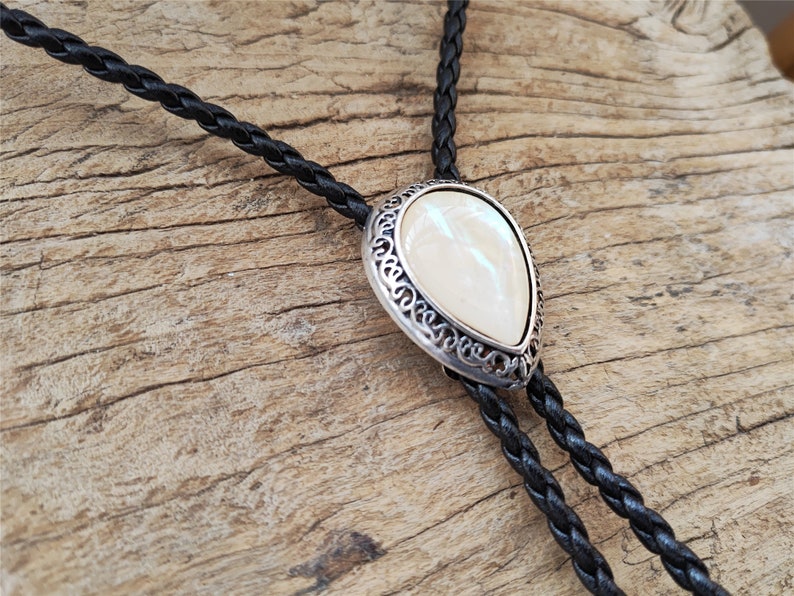 May include: A black braided bolo tie with a silver-toned pendant featuring a white pearlescent stone.