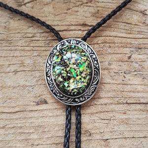 May include: A bolo tie featuring an oval-shaped centerpiece with a black background filled with iridescent green and gold flecks. The centerpiece is encased in a silver-toned metal frame with intricate scrollwork and hangs from a black braided cord.