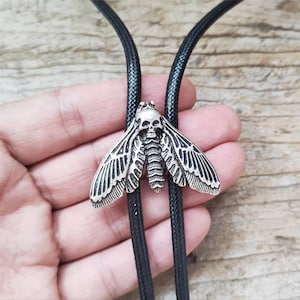 Könnte beinhalten: Silberfarbene Bolo-Krawatte mit einem Motten-Design mit Totenkopf. Die Flügel der Motte sind mit komplizierten Mustern versehen. Die Krawatte hat eine schwarze Kordel. Ein einzigartiges Accessoire.